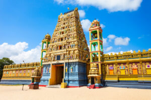 A stunning example of roofs around the world, this colorful Hindu temple features intricate carvings and statues on its tall gopuram tower. Encircled by ornate walls and framed against a bright blue sky with scattered clouds, the sandy ground in front provides a clear and open space.