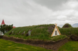 A sod-covered house with a turf roof showcases the unique charm of roofs around the world. Featuring small white-framed windows and a triangular wooden door, the structure sits on a grassy field under a cloudy sky. A red-roofed building is partially visible in the background.