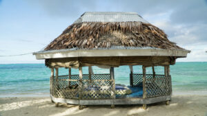 A rustic beachfront hut with a thatched roof, reminiscent of roofs around the world, features wooden lattice sides standing proudly on sandy ground. The turquoise sea sparkles in the background under a partly cloudy sky.