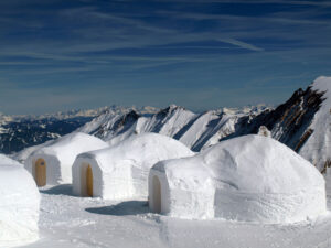 Three snow-covered igloos, showcasing unique roofs from around the world, are set against a backdrop of a mountain range under a blue sky. The igloos have doorways and are surrounded by snowy terrain, with peaks visible in the distance.