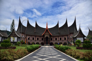 A traditional Minangkabau house, celebrated for its roofs admired around the world, stands under a blue sky. The building features intricate wooden details, nestled within a lush garden with a pathway guiding visitors to the inviting entrance.
