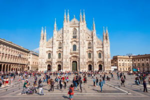 A large crowd gathers in front of the Milan Cathedral, a Gothic architectural marvel with intricate spires and detailed facade, under a clear blue sky. The square is bustling with people, admiring one of the most famous roofs around the world and capturing a lively atmosphere.