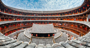 Panoramic view of a traditional Fujian Tulou, showcasing one of the unique roofs around the world. This round, multi-storied earth building features tiered rooftops and wooden balconies beneath a partly cloudy blue sky. Red lanterns hang from the eaves, with clothes visible on the balconies.